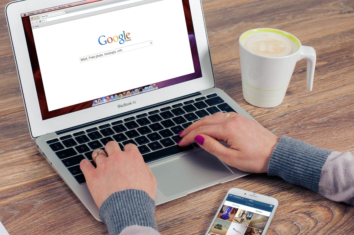 A person with pink nail polish is typing on a MacBook Air with a Google search page open, possibly researching for their digital marketing agency. The desk also has a cup of coffee beside the laptop and a smartphone displaying an image gallery. Opdee Digital Marketing