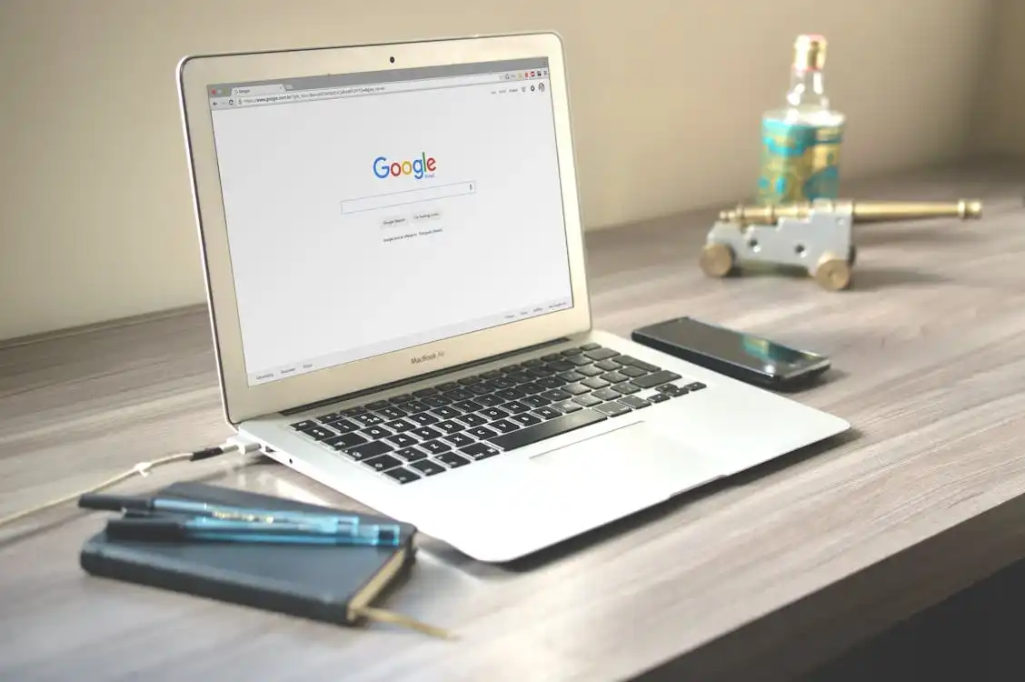 A MacBook displaying the Google search page, highlighting search engine organics, is placed on a wooden desk. Nearby are a smartphone, a notebook with pens, and a decorative glass object next to a small metallic cannon figure. The scene is well-lit with a neutral, beige-colored background. Opdee Digital Marketing