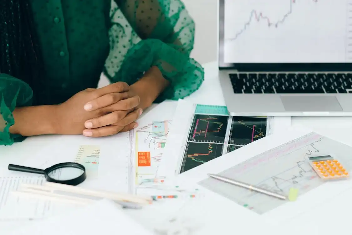 Person wearing a green blouse sits at a desk filled with various financial documents, charts, magnifying glass, calculator, ruler, pencil, and an open laptop displaying a stock market graph. The person's hands are folded and resting on the desk, reminiscent of someone preparing for a meeting at a digital marketing agency. Opdee Digital Marketing