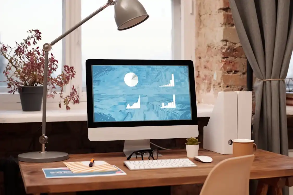 A home office setup with a modern desktop computer displaying charts and graphs, hinting at a busy digital agency. The desk has a keyboard, mouse, notepad, pen, glasses, a small potted plant, and white file organizers. A desk lamp is turned on, and there's a window with potted plants behind it. Opdee Digital Marketing
