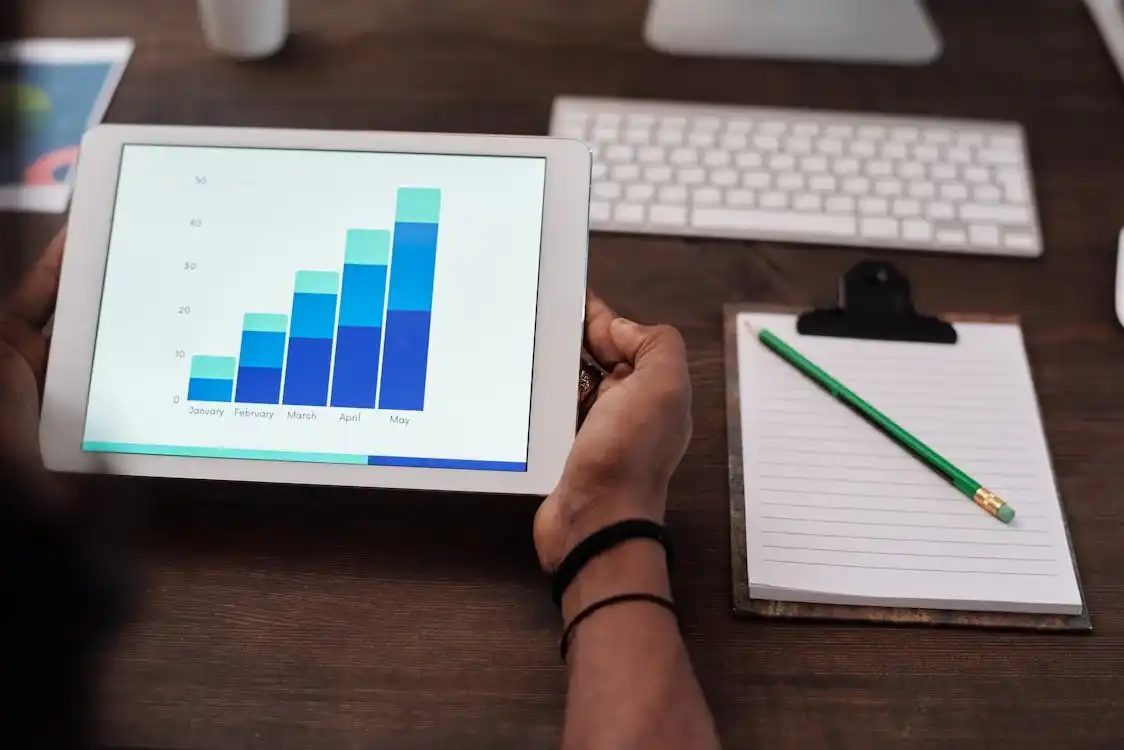 A person holds a tablet displaying a bar chart with monthly data from January to May. A white keyboard, a clipboard with a blank notepad, and a green pencil are placed on the wooden desk in the background, possibly highlighting trends tracked by their digital agency. Opdee Digital Marketing