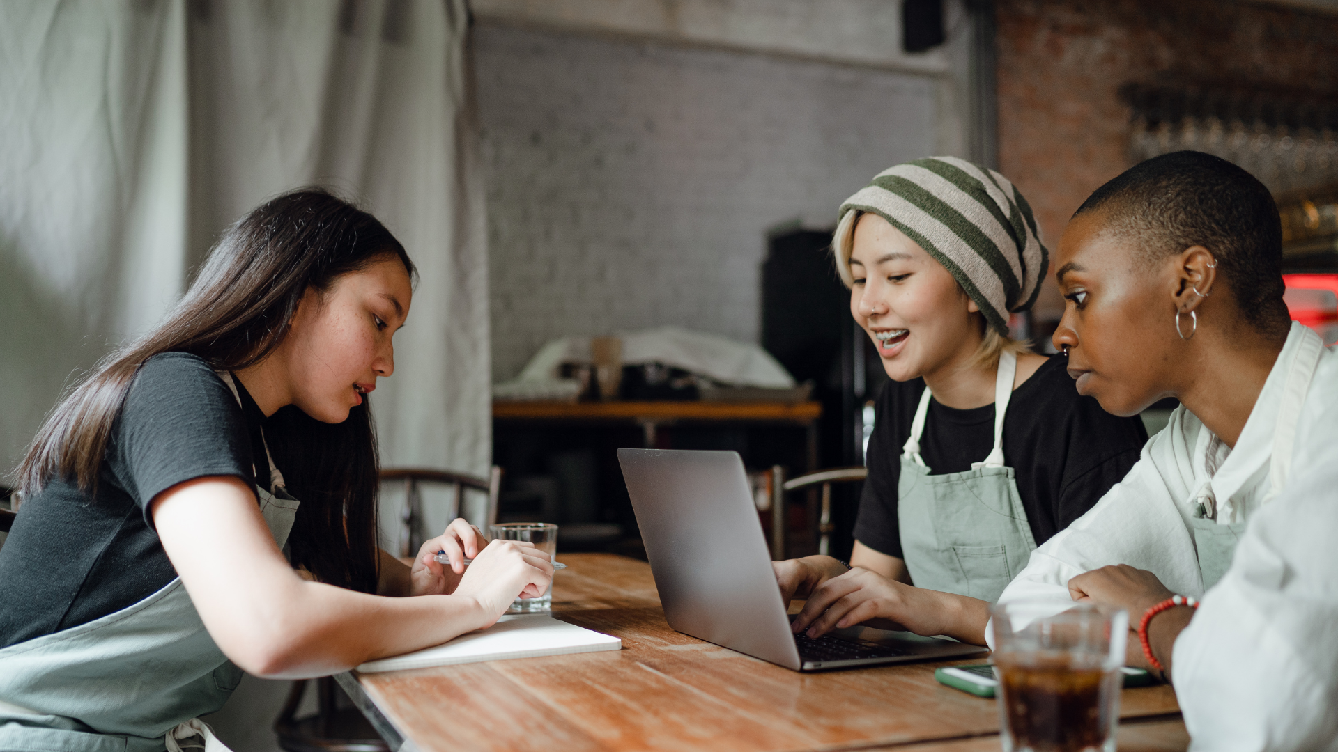 Three people wearing aprons sit at a wooden table, collaborating on their social media strategy. One writes in a notebook, another types and smiles at a laptop, while the third looks on. Drinks sit nearby in this cozy indoor setting. Opdee Digital Marketing