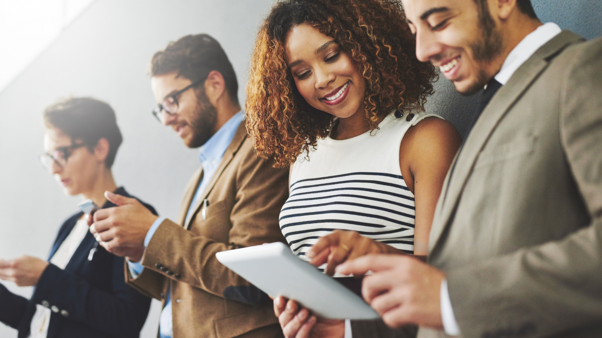 Four professionally dressed people stand in a row, smiling and interacting with digital devices, showcasing teamwork essential for scaling social media or executing a successful social media strategy against a plain wall. Opdee Digital Marketing