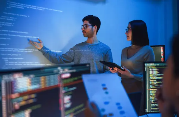 Two people stand before a projected screen displaying code, engaged in discussion about AI workshops. Computer monitors with code in the foreground highlight a focused tech work environment. Opdee Digital Marketing