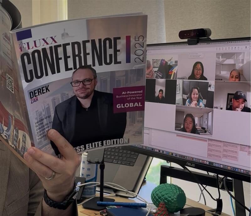 A person holds a magazine titled "Conference 2025" in front of a computer showing a video call with six people. The desk has a keyboard, mouse, wires, and a green 3D-printed object. Opdee Digital Marketing