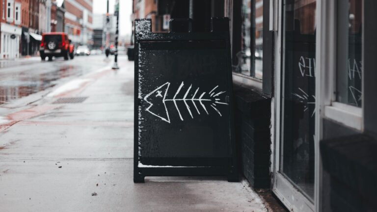A black sandwich board with a white chalk drawing of a fish skeleton stands on a wet sidewalk outside a building, subtly hinting at brand guideline mistakes in an urban area on a rainy day. Opdee Digital Marketing