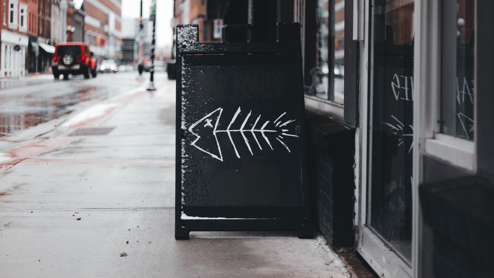 A black sandwich board with a white chalk drawing of a fish skeleton stands on a wet sidewalk outside a building, subtly hinting at brand guideline mistakes in an urban area on a rainy day. Opdee Digital Marketing