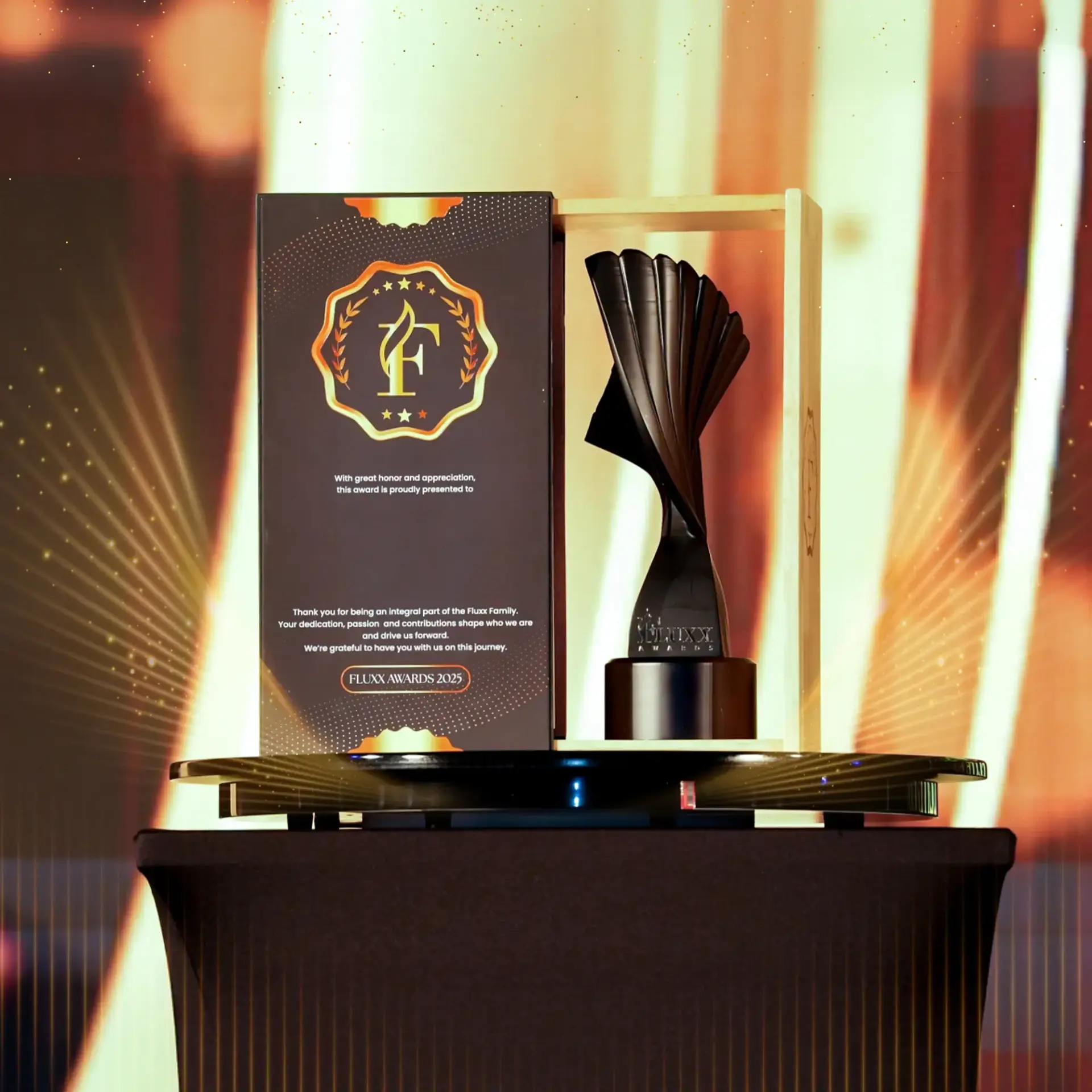 A black sculptural award trophy stands next to a decorative plaque with gold text and accents, both displayed on a black round pedestal against a blurred, warm-toned background. Opdee Digital Marketing