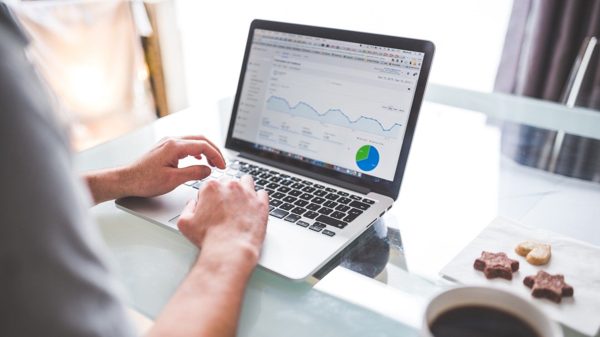 A person types on a laptop displaying charts and graphs, including a line graph and pie chart analyzing Google Ads performance, on a glass table with coffee and cookies nearby. Opdee Digital Marketing