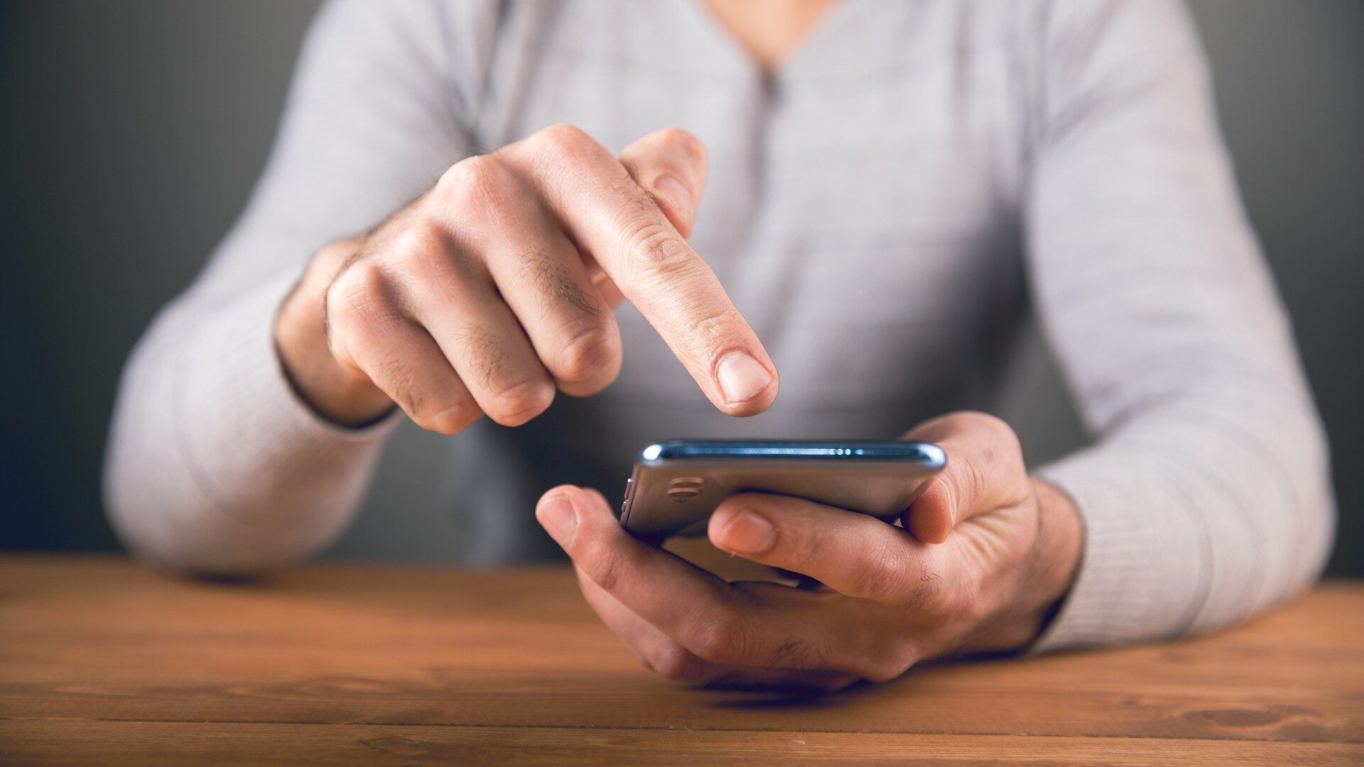 A person wearing a light gray long-sleeve shirt is using a smartphone, possibly researching their customer buying journey, tapping the screen with one hand while sitting at a wooden table. Opdee Digital Marketing